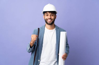 Young architect Moroccan man with helmet and holding blueprints over isolated background celebrating a victory in winner position