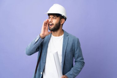 Young architect Moroccan man with helmet and holding blueprints over isolated background shouting with mouth wide open to the side