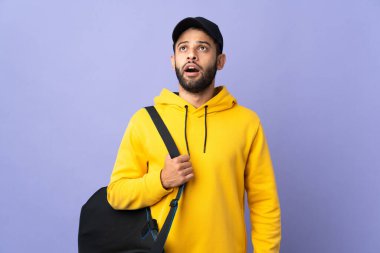 Young sport Moroccan man with sport bag isolated on purple background looking up and with surprised expression