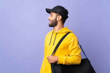 Young sport Moroccan man with sport bag isolated on purple background laughing in lateral position