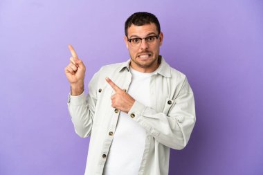 Brazilian man over isolated purple background frightened and pointing to the side