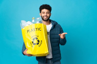 Young Moroccan man holding a bag full of plastic bottles to recycle over isolated background pointing finger to the side