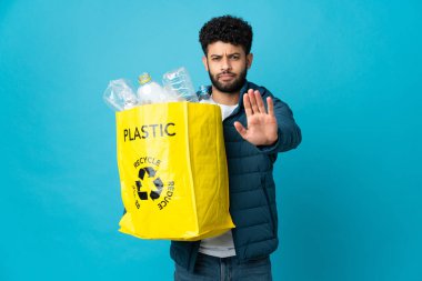 Young Moroccan man holding a bag full of plastic bottles to recycle over isolated background making stop gesture