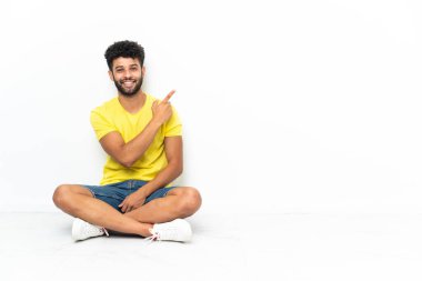 Young Moroccan handsome man sitting on the floor over isolated background pointing finger to the side