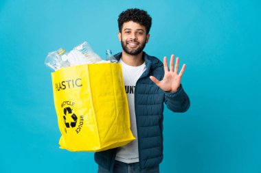 Young Moroccan man holding a bag full of plastic bottles to recycle over isolated background counting five with fingers