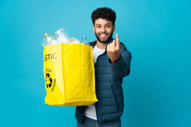 Young Moroccan man holding a bag full of plastic bottles to recycle over isolated background doing coming gesture