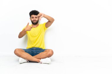 Young Moroccan handsome man sitting on the floor over isolated background focusing face. Framing symbol