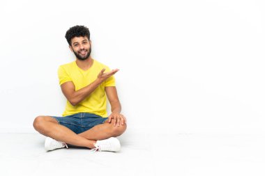Young Moroccan handsome man sitting on the floor over isolated background presenting an idea while looking smiling towards