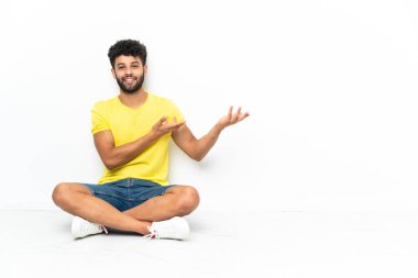 Young Moroccan handsome man sitting on the floor over isolated background extending hands to the side for inviting to come