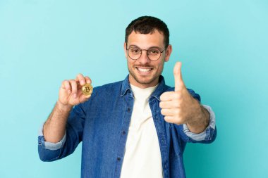 Brazilian man holding a Bitcoin over isolated blue background with thumbs up because something good has happened