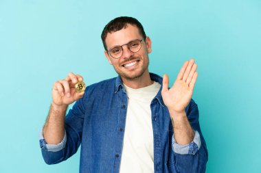 Brazilian man holding a Bitcoin over isolated blue background saluting with hand with happy expression