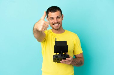 Brazilian man holding a drone remote control over isolated blue background with thumbs up because something good has happened