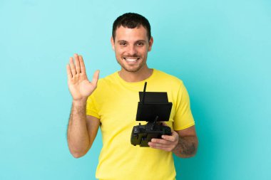 Brazilian man holding a drone remote control over isolated blue background saluting with hand with happy expression