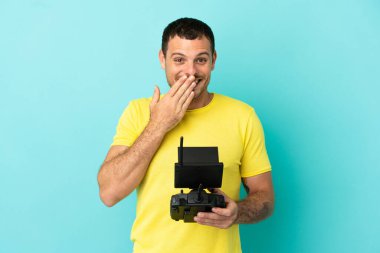 Brazilian man holding a drone remote control over isolated blue background happy and smiling covering mouth with hand