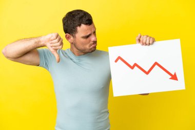 Brazilian man over isolated purple background holding a sign with a decreasing statistics arrow symbol and doing bad signal