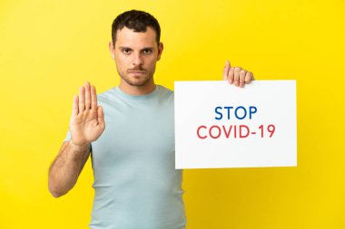 Brazilian man over isolated purple background holding a placard with text Stop Covid 19 and doing stop sign