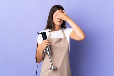 Brazilian woman using hand blender isolated on purple background covering eyes by hands. Do not want to see something
