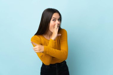 Teenager girl isolated on blue background pointing to the side to present a product and whispering something