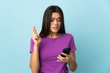 Teenager girl isolated on pink background using mobile phone with fingers crossing