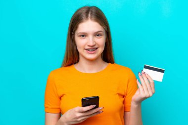 Genç Rus kız, mavi arka planda izole edilmiş cep telefonuyla kredi kartı satın alıyor.