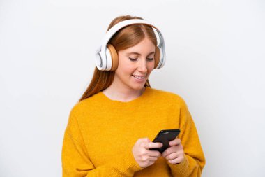 Genç kızıl saçlı kadın beyaz arka planda izole edilmiş müzik dinliyor ve cep telefonu arıyor.