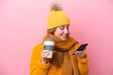 Kışlık ceket giyen genç kızıl saçlı kadın pembe arka planda izole edilmiş. Elinde kahve ve cep telefonu var.