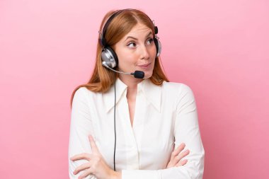 Tele pazarlamacı kızıl saçlı kadın pembe arka planda izole edilmiş bir kulaklıkla çalışıyor. Omuzlarını kaldırırken şüpheli hareketler yapıyor.