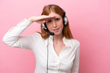 Tele pazarlamacı kızıl saçlı kadın pembe arka planda izole edilmiş bir kulaklıkla çalışıyor. Bir şeye bakmak için çok uzaklara bakıyor.