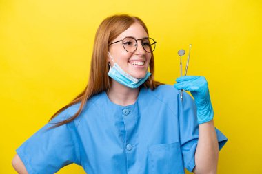 Genç kızıl saçlı dişçi kadın sarı arka planda izole edilmiş. Kolları kalçasında ve gülümsüyor.