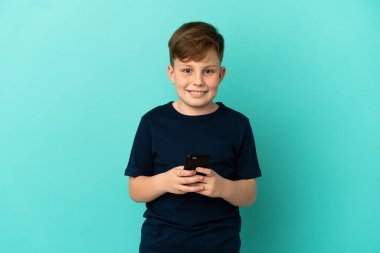 Mavi arka planda izole edilmiş küçük kızıl çocuk cep telefonuyla mesaj gönderiyor.