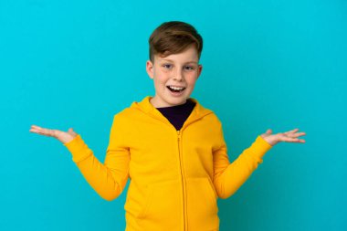 Küçük kızıl saçlı çocuk mavi arka planda izole edilmiş ve şok olmuş bir yüz ifadesi var.