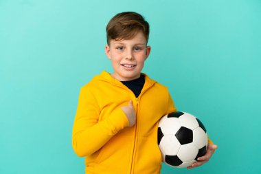 Mavi arka planda futbol oynayan kızıl saçlı çocuk şaşırmış yüz ifadesi ile izole edilmiş.