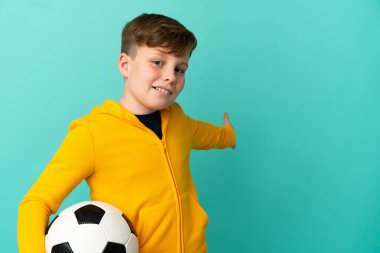 Kızıl saçlı çocuk mavi arka planda izole bir şekilde futbol oynuyor. Davet etmek için ellerini yana açıyor.