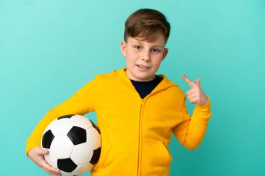 Kırmızı saçlı çocuk mavi arka planda izole bir şekilde futbol oynuyor.