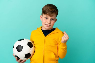 Mavi arka planda izole bir şekilde futbol oynayan kızıl saçlı çocuk para hareketi yapıyor.