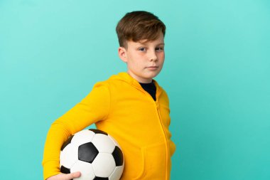 Küçük kızıl saçlı çocuk mavi arka planda futbol topuyla izole edilmiş.