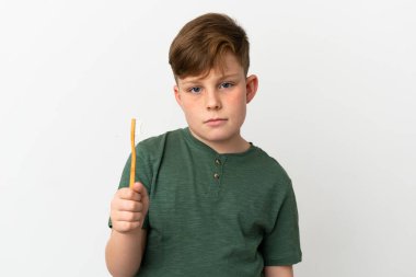 Küçük kızıl saçlı çocuk, elinde diş fırçası olan üzgün bir ifadeyle beyaz arka planda izole edilmiş.