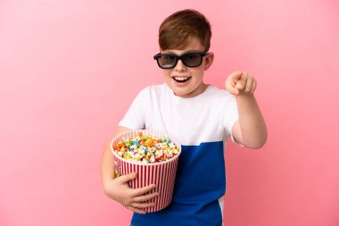 Küçük kızıl saçlı çocuk pembe arka planda gözlüklü ve elinde büyük bir kova patlamış mısırla ön tarafı işaret ediyor.