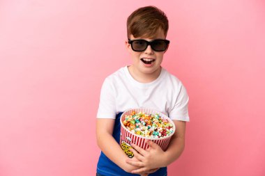 Küçük kızıl saçlı çocuk pembe arka planda 3 boyutlu gözlüklü ve elinde patlamış mısır dolu bir kova tutuyor.