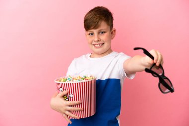 Küçük kızıl saçlı çocuk pembe arka planda 3 boyutlu gözlüklü ve elinde patlamış mısır dolu bir kova tutuyor.