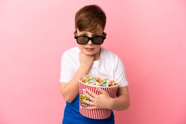 Küçük kızıl saçlı çocuk pembe arka planda 3 boyutlu gözlüklü ve elinde patlamış mısır dolu bir kova tutuyor.