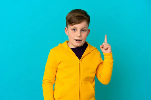 指を持ち上げながら解決策を実現しようとしている青い背景に隔離された小さな赤毛の少年 — ストック写真