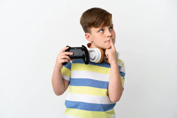 一个红头发的小男孩拿着一个白色背景的游戏垫 一边抬头一边怀疑 — 图库照片