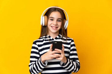 Küçük kız sarı arka planda izole edilmiş. Cep telefonuyla müzik dinliyor ve ön tarafa bakıyor.