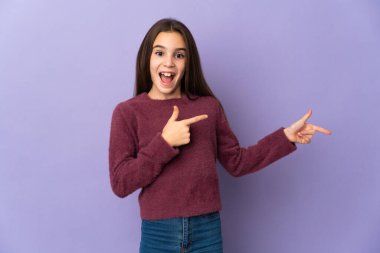Mor arka planda izole edilmiş küçük kız şaşırmış ve işaret ediyor.
