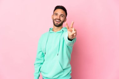 Pembe arka planda izole edilmiş genç beyaz adam gülümsüyor ve zafer işareti gösteriyor.