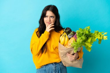 Elinde alışveriş çantası olan genç bir kadın mavi arka planda izole edilmiş bir şekilde yukarı bakarken şüpheleri var.