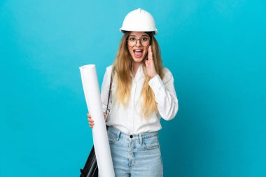 Miğferli ve elinde mavi arka planda izole edilmiş, şaşırmış ve şok olmuş yüz ifadesi olan genç mimar kadın.