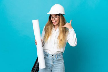 Miğferli ve elinde mavi arka planda izole edilmiş planları olan genç bir mimar kadın telefon jesti yapıyor. Bana geri dönüş işareti ver.