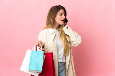 Alışveriş çantalı genç bir kadın pembe arka planda izole edilmiş alışveriş torbaları tutuyor ve cep telefonuyla arkadaşını arıyor.